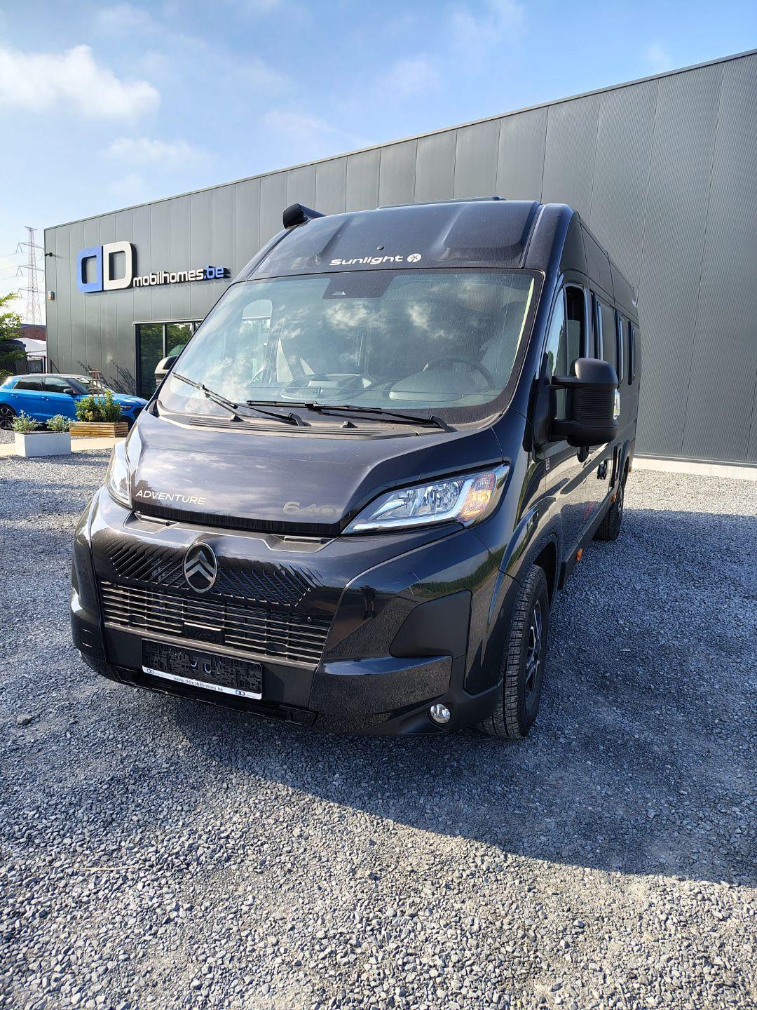 Front three-quarter view of a black Sunlight motorhome van on a gravel lot, with a gray industrial building and 'mobilhomes.de' sign in the background.