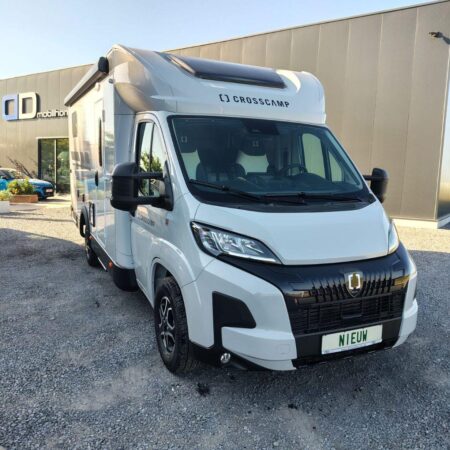 White Crosscamp camper van parked on a gravel lot outside a modern building, shows solar panel on roof and large front grille with badge, Dutch license plate reading NIEUW in green letters
