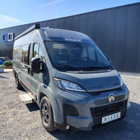 Gray camper van labeled 'Adventure 640' parked on a gravel lot beside a dark metal building, sunny day.