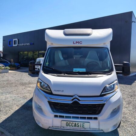 Front view of a white camper van with an overcab berth, parked on a gravel lot in front of a showroom with 'mobilhomes.be' signage.