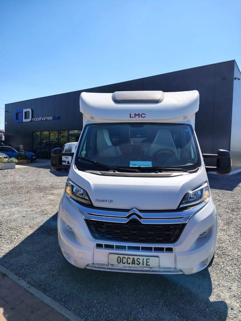Front view of a white camper van with an overcab berth, parked on a gravel lot in front of a showroom with 'mobilhomes.be' signage.