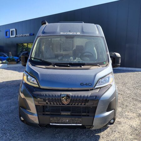 Front view of a gray Peugeot camper van (model 640) with Sunlight branding, parked in a lot beside a dark building with 'mobilhomes.be' sign.