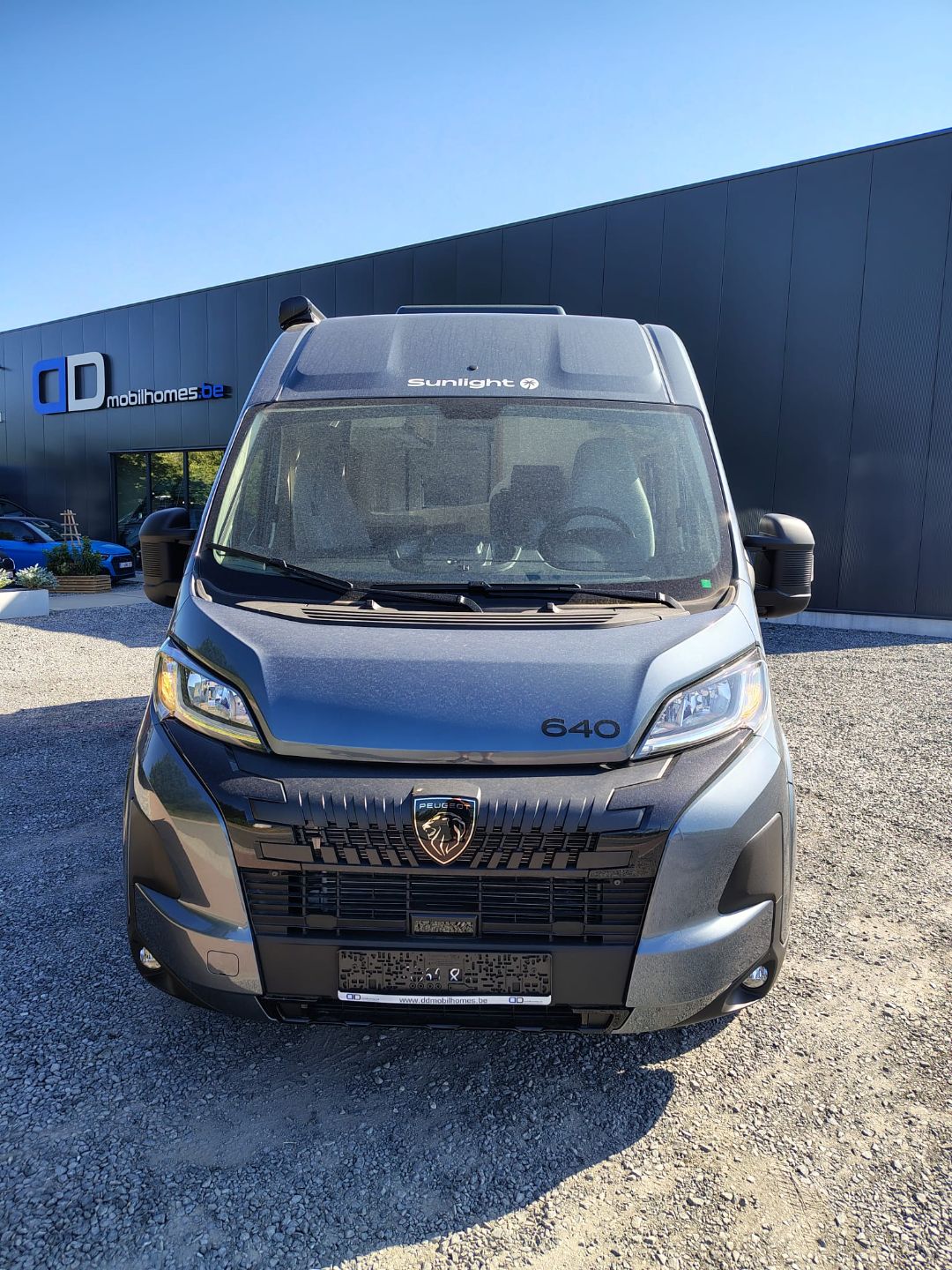 Front view of a gray Peugeot camper van (model 640) with Sunlight branding, parked in a lot beside a dark building with 'mobilhomes.be' sign.