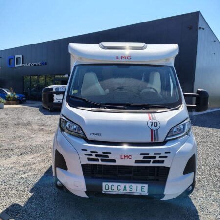 Front view of a white LMC motorhome parked in a gravel lot outside a showroom with a 'CD mobilhomes' sign in the background.