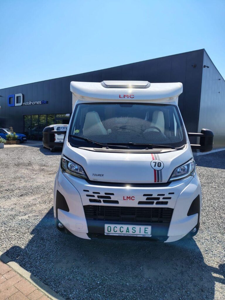 Front view of a white LMC motorhome parked in a gravel lot outside a showroom with a 'CD mobilhomes' sign in the background.