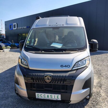 Front view of a silver Peugeot camper van with '640' badge, parked on gravel outside a dark showroom building with 'mobilhomes.de' signage in the background, sunny sky above.