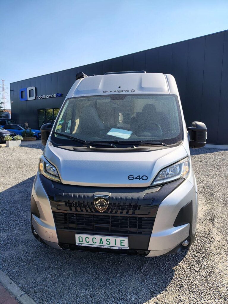 Front view of a silver Peugeot camper van with '640' badge, parked on gravel outside a dark showroom building with 'mobilhomes.de' signage in the background, sunny sky above.