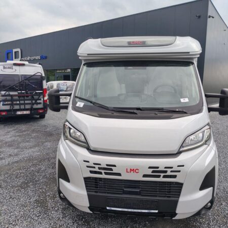 Front view of a white LMC motorhome parked in a lot beside a dark industrial building with other RVs nearby.