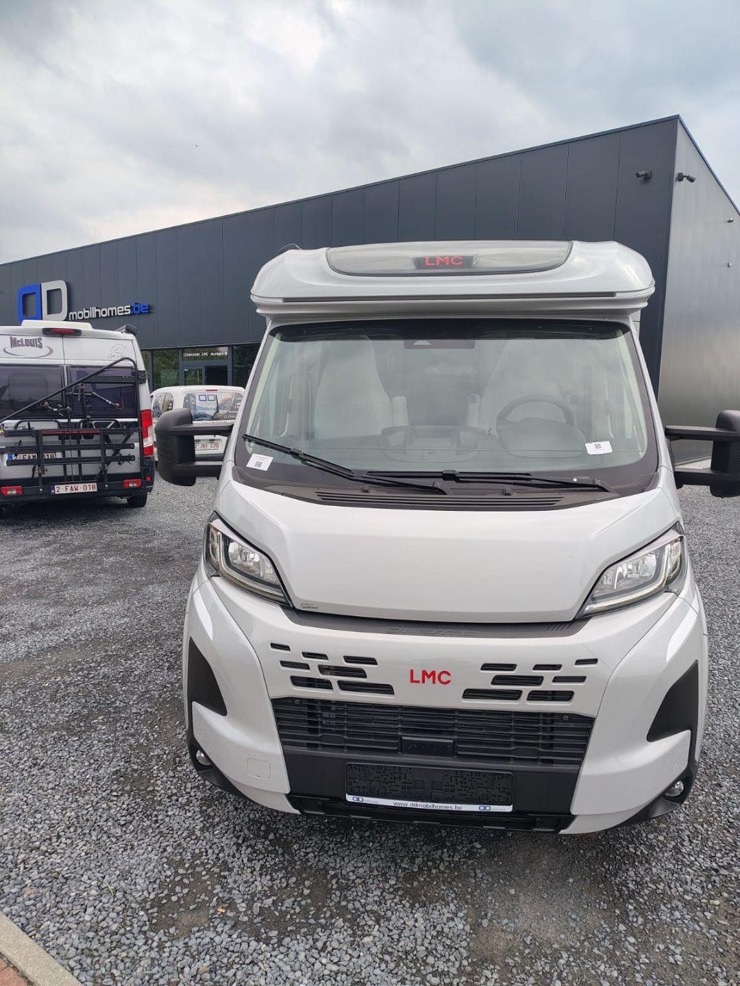 Front view of a white LMC motorhome parked in a lot beside a dark industrial building with other RVs nearby.