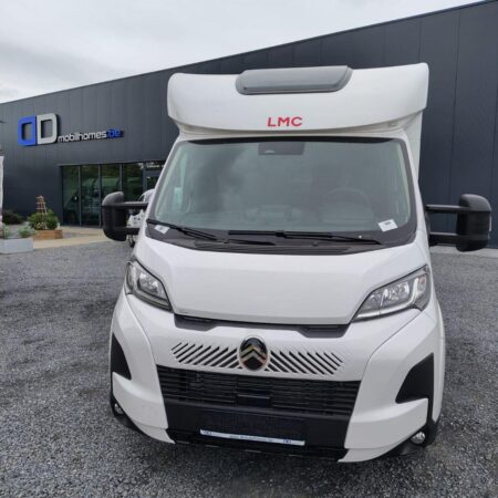 Front view of a white LMC camper van with a large over-cab section, parked on gravel outside a modern dealership with dark gray panels.