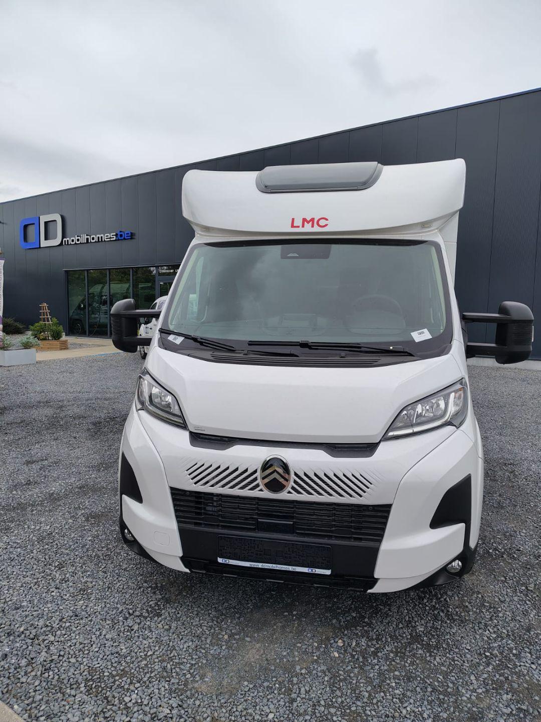 Front view of a white LMC camper van with a large over-cab section, parked on gravel outside a modern dealership with dark gray panels.